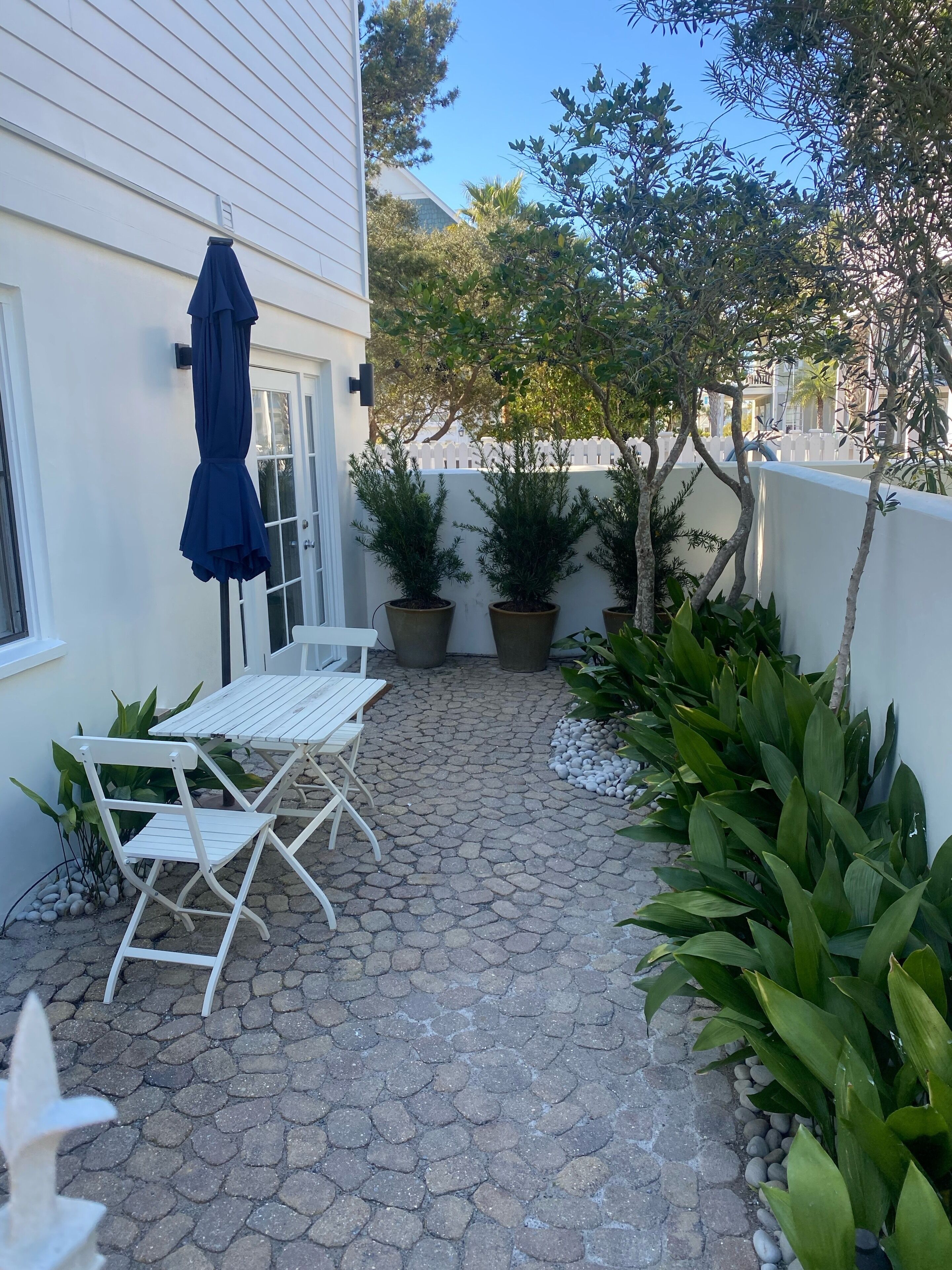 Un patio con una mesa y sillas blancas, una sombrilla azul y plantas en macetas.