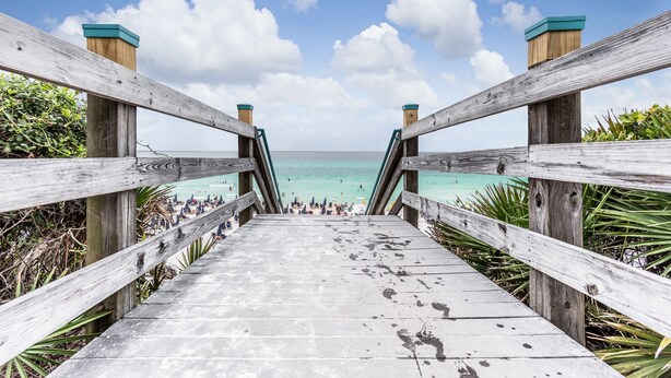 A wooden boardwalk leading to a beach with clear turquoise water and a clear sky.