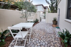 Un patio con una mesa y sillas blancas, rodeado de plantas en macetas y un sendero de piedra que lleva a una reja.