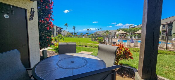 A patio area with a round table and chairs, a view of a swimming pool, and a lawn with tropical plants. - Incredible Ocean View,  Most Desirable Unit in the Complex (Kihei)