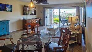 A dining area with a glass table and chairs, a blue cabinet, a television, and a view of the outdoors through a sliding door. - Incredible Ocean View,  Most Desirable Unit in the Complex (Kihei)