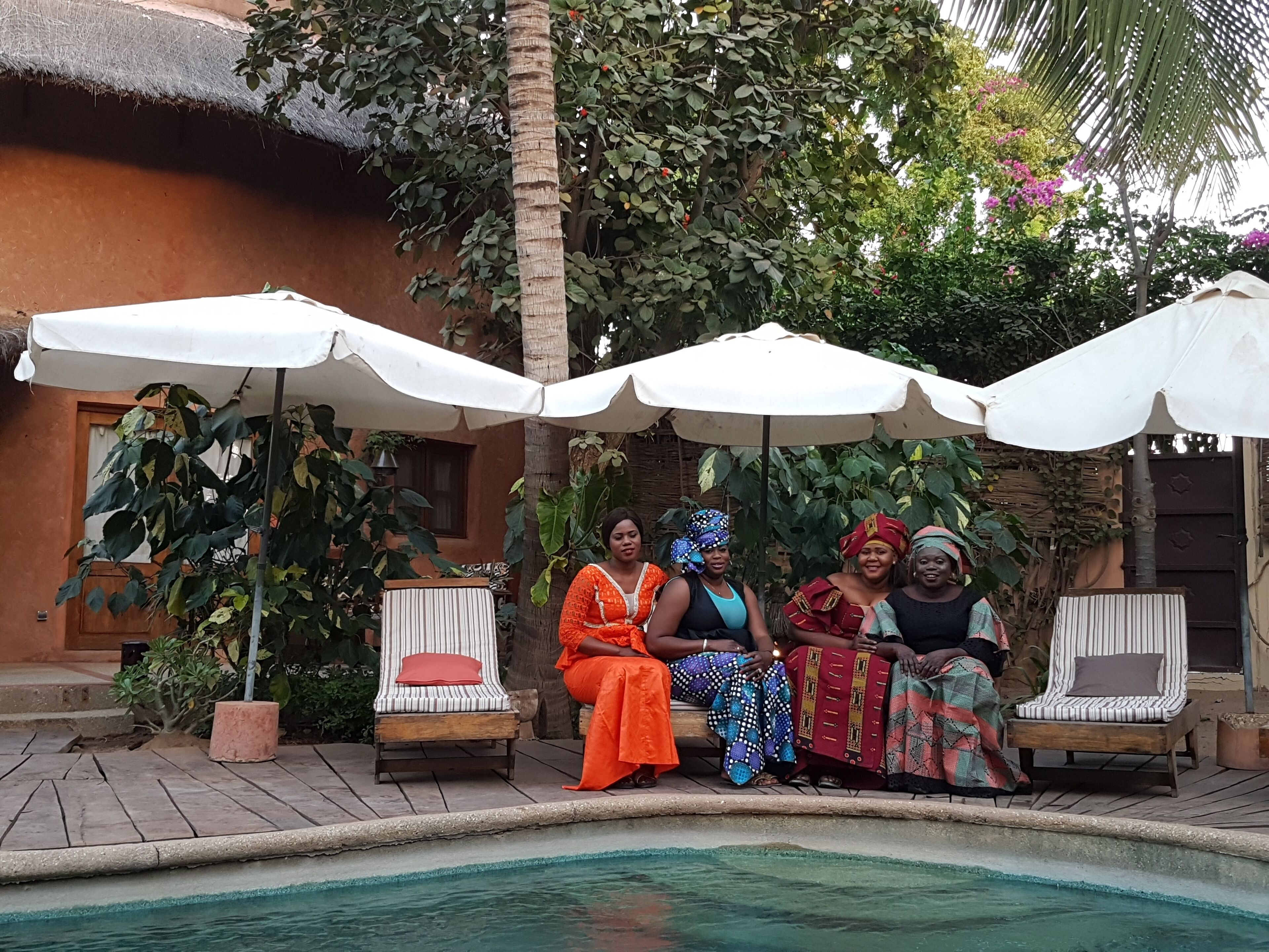 A poolside area with four lounge chairs under white umbrellas, surrounded by lush greenery and a thatched-roof building.