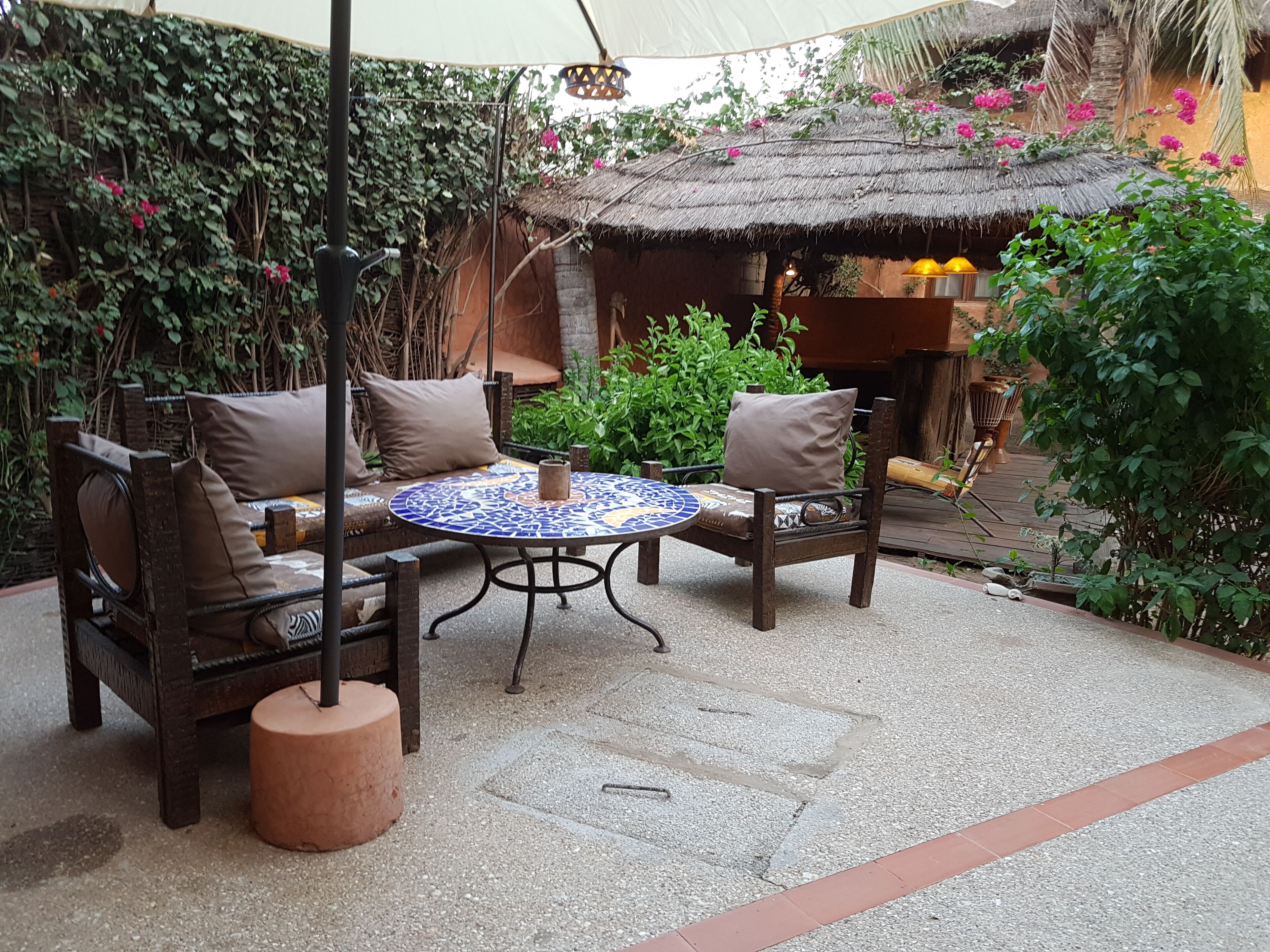 An outdoor seating area with a round table and two chairs, surrounded by a garden with a thatched-roof structure.