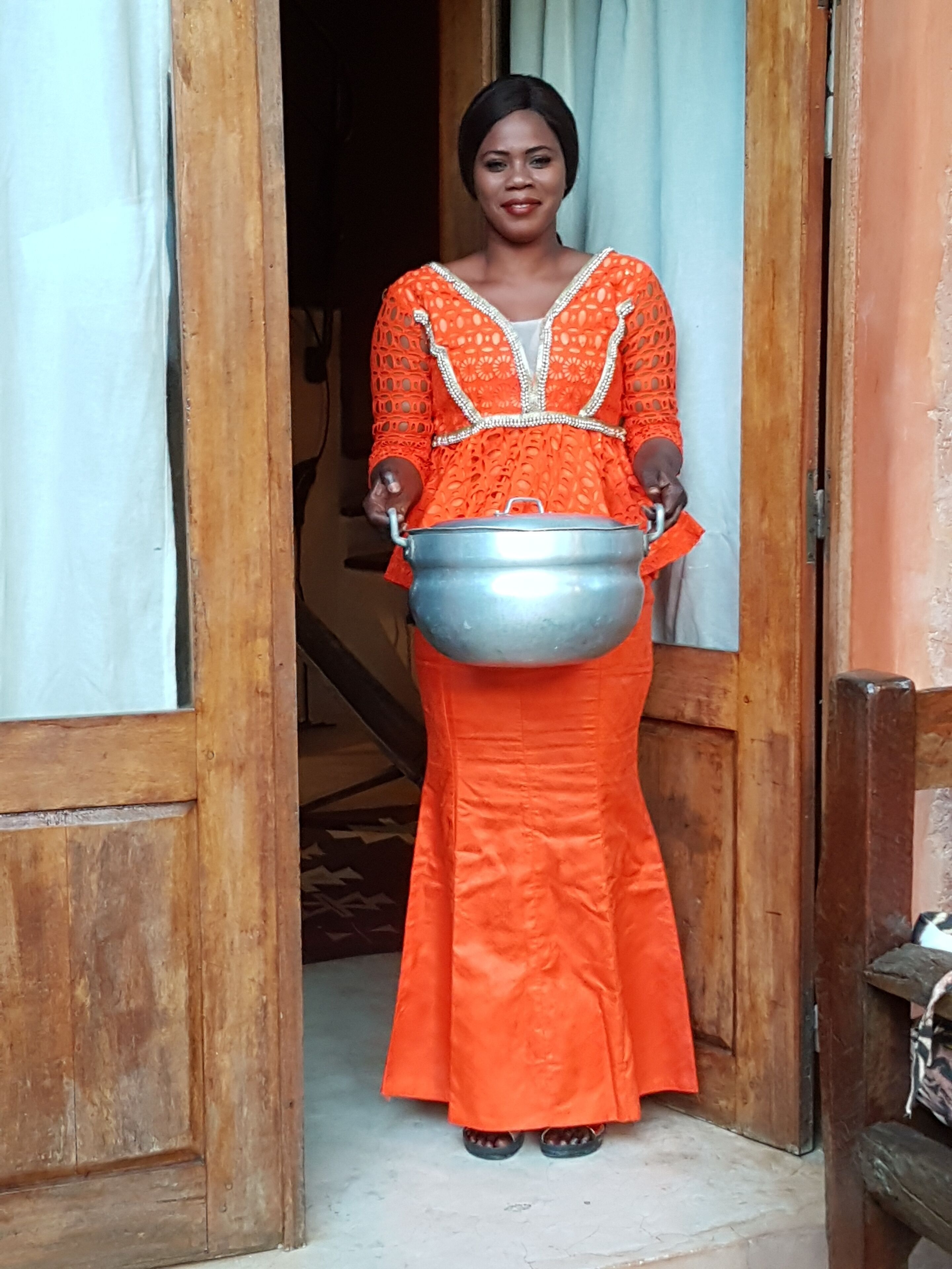 Una persona con un vestido naranja sostiene una olla de metal, de pie frente a la entrada de una puerta de madera.