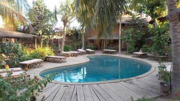 A pool area with wooden deck, lounge chairs, and tropical plants.