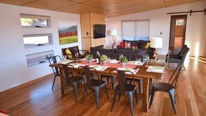 Un coin repas avec une table en bois dressée pour six personnes, entourée de chaises en métal. On y trouve également un canapé et un téléviseur fixé au mur.
