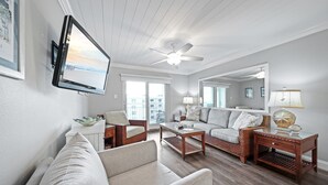 A living room with a flat-screen TV mounted on the wall, a ceiling fan, wicker furniture, and a large window offering a view of the city. - Top-floor ocean view 2/2 with a balcony, heated beachfront pool, and beach access (New Smyrna Beach)
