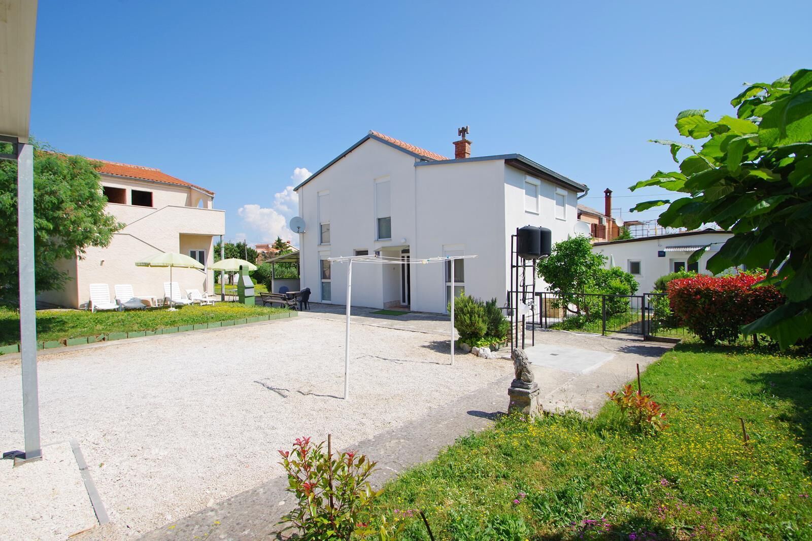 A residential area with a white building, a gravel driveway, and a small garden with flowers and shrubs.