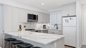 A modern kitchen with white cabinets, a marble countertop, a built-in microwave, a refrigerator, and bar stools. - Newly renovated townhouse - bike right to your door, Private hot tub, BBQ (Silver Star Mountain)