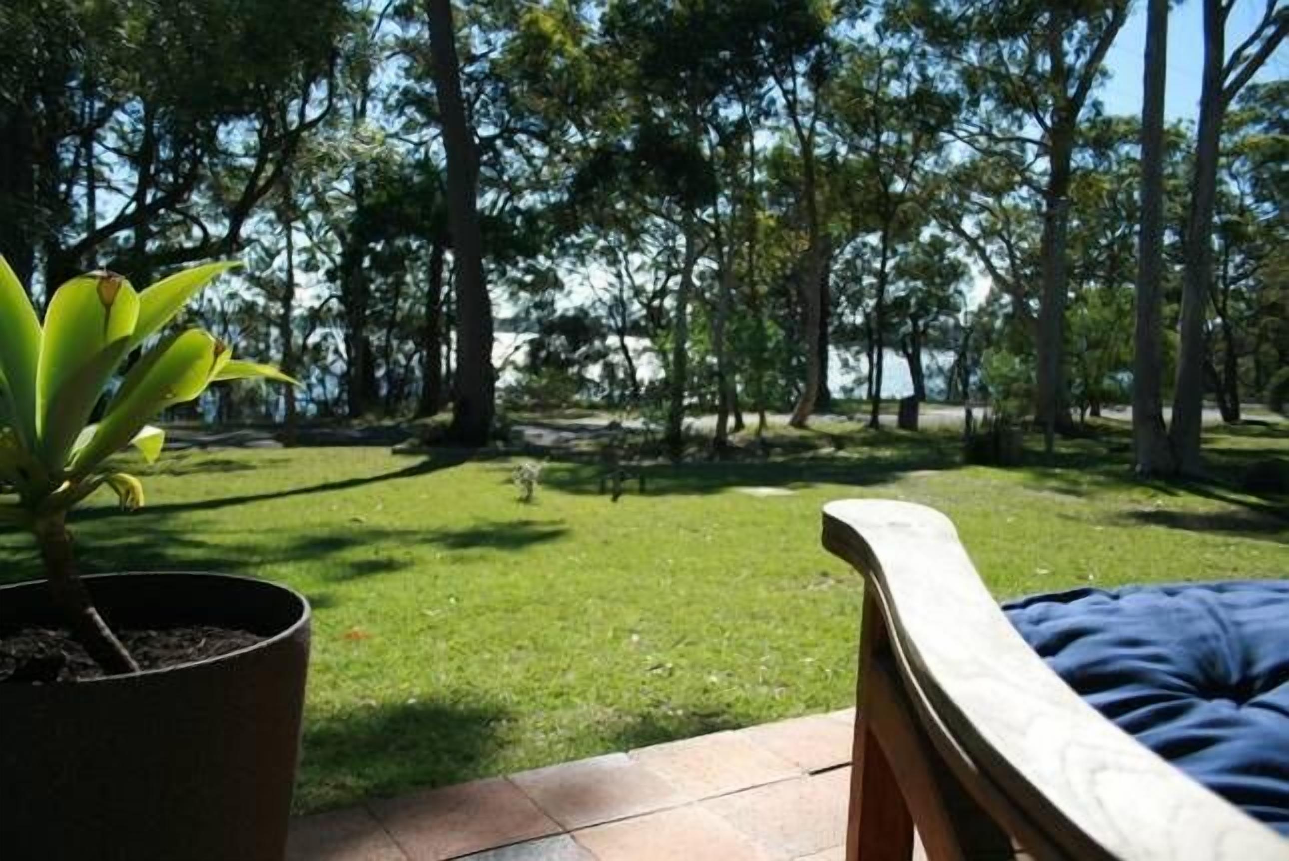 A patio area with a view of a grassy lawn and trees, a potted plant, and a wooden bench.