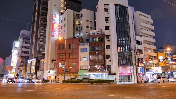 Un edificio de varios pisos con diversos comercios en la planta baja, incluyendo un restaurante bien iluminado y una tienda de conveniencia.
