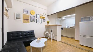 Una sala de estar moderna con un sofá negro y una mesa de centro blanca, una cocina con microondas y refrigerador, y una pared decorada con cuadros y un reloj.