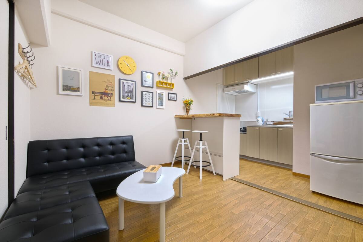 A modern living room with a black sofa and a white coffee table, a kitchen with a microwave and refrigerator, and a wall decorated with framed pictures and a clock.
