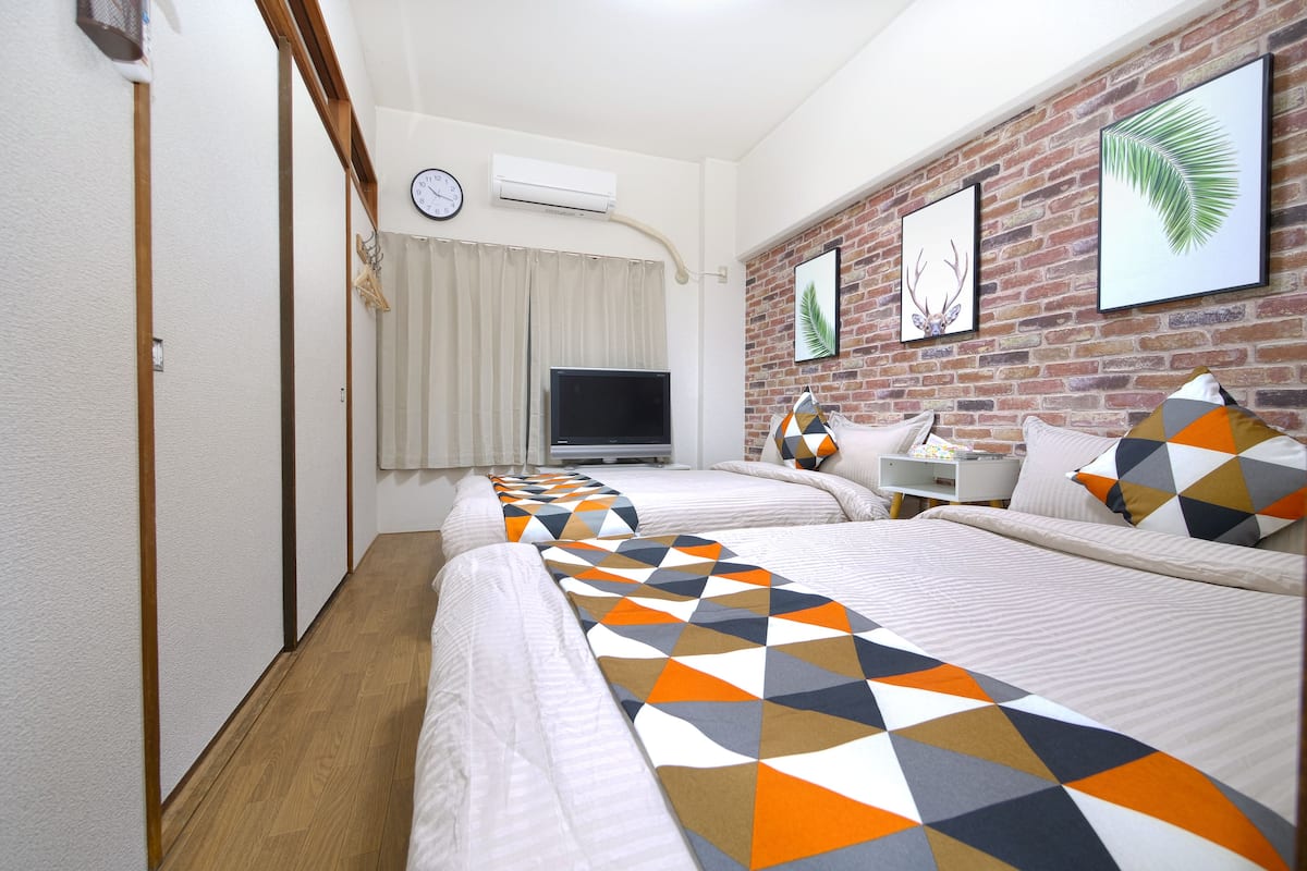 A hotel room with a brick wall, a television, a desk with a chair, and two beds with patterned bedding.