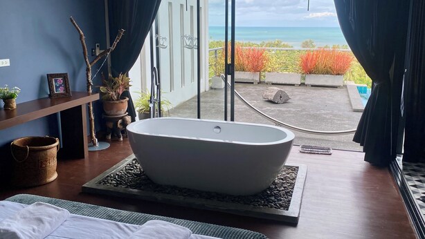 Un bagno moderno con una vasca da bagno indipendente, un'ampia finestra con tende e vista sull'oceano e sulla vegetazione circostante.