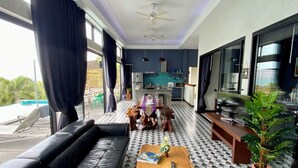 Un soggiorno moderno con un divano in pelle nera, un tavolino in legno e una televisione. Sono presenti dei ventilatori a soffitto, un'area cucina sullo sfondo e un'ampia finestra con tende.