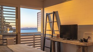 A bedroom with a bed, a television on a wooden stand, and a view of the sea through a window.