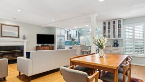 A modern living room with a fireplace, a dining area with a wooden table and chairs, and a kitchen in the background. - Private Oasis with a Large Saltwater Pool Close to Everything - Screened Porch and Deck Open Concept (Folly Beach)
