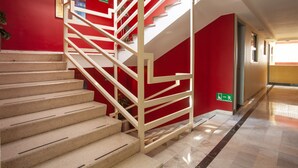 A staircase with white railings and beige steps, leading to a landing with a red wall and a green exit sign.