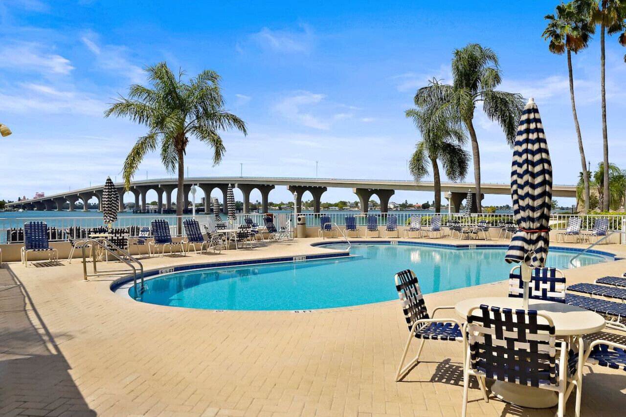 A poolside area with lounge chairs and umbrellas, palm trees, and a bridge in the background.