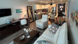 Un salon moderne avec un canapé blanc, une table basse et un téléviseur. Au fond, on aperçoit une cuisine avec un coin repas.