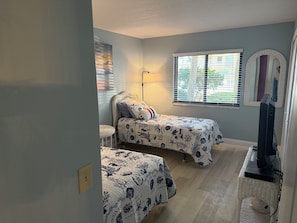Une chambre avec deux lits, une fenêtre avec des stores, une lampe, un miroir et un bureau avec un ordinateur.