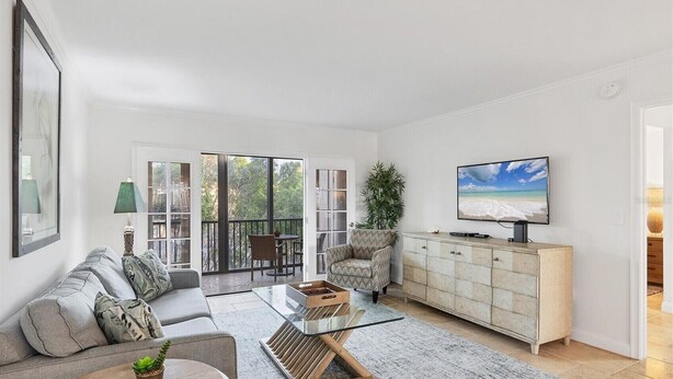 A spacious living room with a sofa, coffee table, TV, and a balcony with a view of the outdoors. - Beautifully Updated & Decorated Gulf Side Garden View Condo (Siesta Key)