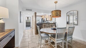 A dining area with a round table, chairs, a hanging light fixture, and a kitchen in the background. - Beautifully Updated & Decorated Gulf Side Garden View Condo (Siesta Key)
