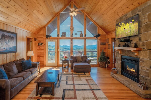 L’intérieur d’un chalet en rondins, avec une cheminée, des meubles en bois, un téléviseur et une vue sur l’extérieur.
