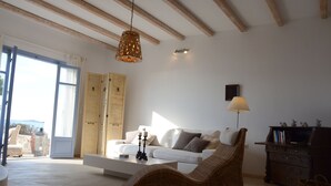 Ein geräumiges Wohnzimmer mit einem weißen Sofa, einem Korbstuhl, einem Holzschrank und einem großen Fenster mit Vorhängen.