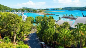 A coastal road leading to a marina with boats, surrounded by lush greenery and hills in the background.