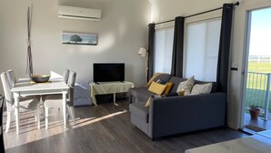 Ein modernes Wohnzimmer mit grauem Sofa, einem Esstisch mit Stühlen, einem Flachbildfernseher und einem großen Fenster mit Vorhängen.