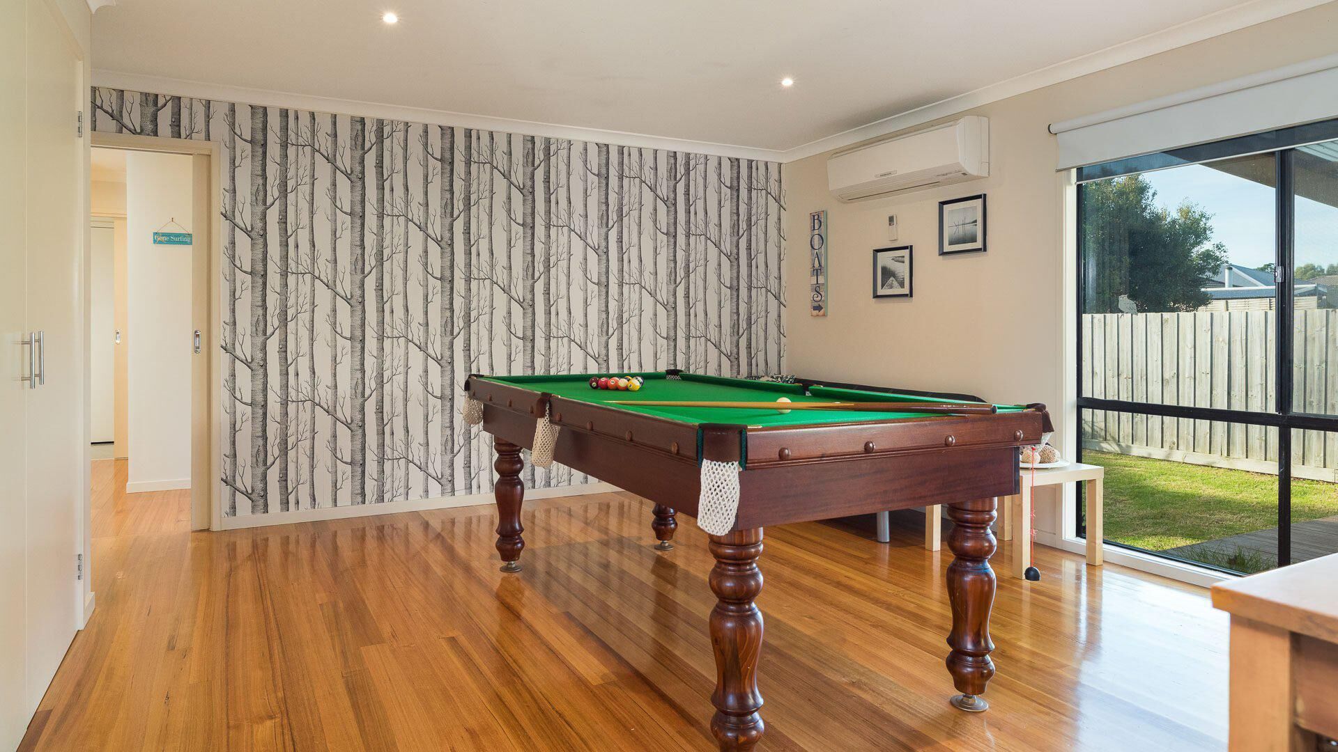 A pool table in a room with wooden flooring, a wall with a tree pattern, and a large window with a view of a backyard.