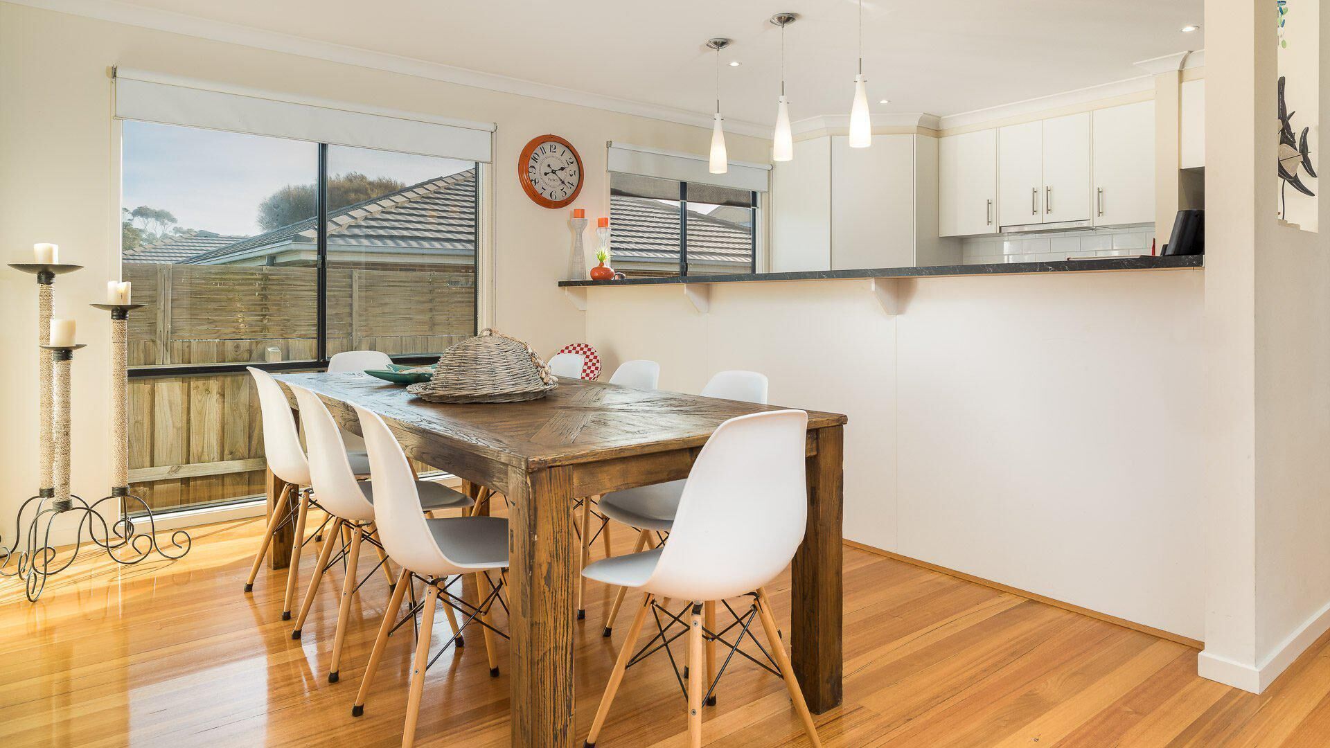 Un espace repas comprenant une table en bois, des chaises blanches, une horloge, des luminaires suspendus et une vue à travers une grande fenêtre.