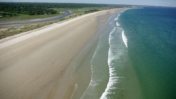 Eine Vogelperspektive zeigt einen Strand mit einem breiten Sandstreifen, einem klaren blauen Ozean und einer Straße, die parallel zum Strand verläuft.
