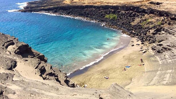 A coastal landscape with clear blue waters, a sandy beach, and rocky terrain.