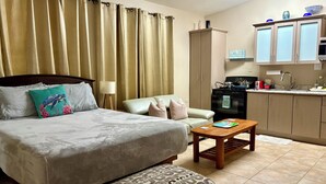 Hotelkamer met een bed, een slaapbank, een gestreept voetenbankje, een kleine tafel, een keuken met kasten en apparatuur, en een raam met gordijnen.