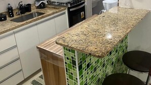 A kitchen with a granite countertop, a stove, a sink, and a green-tiled backsplash. There are two chairs and a white trash bin. - Apt-207 in Comfortable Condominium 350m from the beach W/Air cond (Praia Grande)