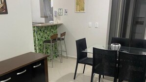 A dining area with a glass table and black chairs, a small kitchen with green tiles, and a white cabinet with drawers. - Apt-207 in Comfortable Condominium 350m from the beach W/Air cond (Praia Grande)