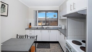 Een functionele keuken met witte kastjes, een raam, een spoelbak, een kookplaat en een eethoek met een tafel en stoelen.