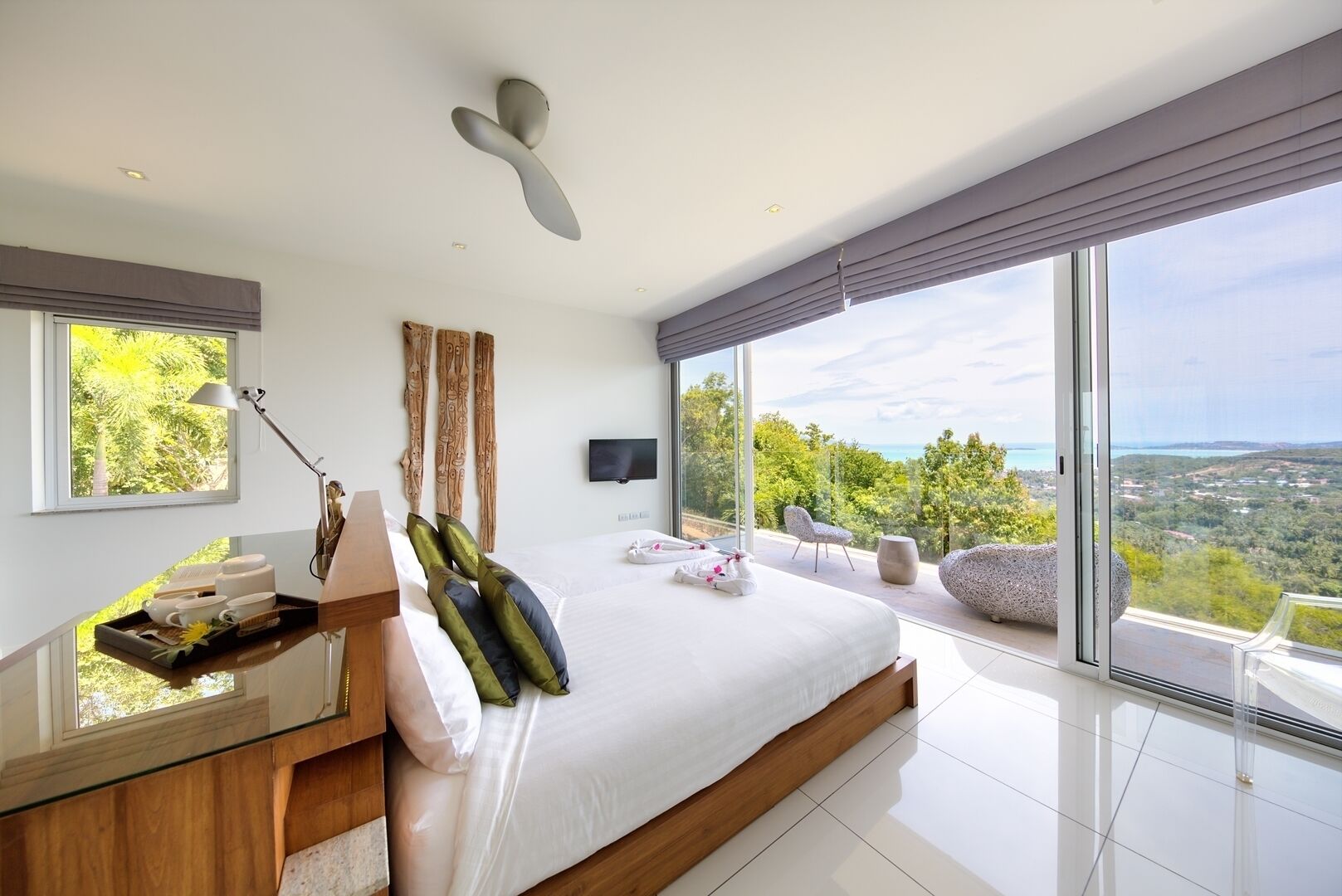 A modern bedroom with a large bed, a bedside table with a lamp, a TV on the wall, and a view of the landscape through large windows.