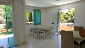 A spacious room with a dining table and chairs, a sofa, and a sliding glass door leading to a balcony with a view of greenery. - BLACK FRIDAY FEBRUARY. Beautiful duplex house, Ilhéus-Bahia, Olivença, Cond.  (Ilheus)