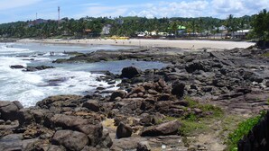 A rocky beach with waves crashing onto the shore, a sandy beach in the background, and a line of trees and buildings. - BLACK FRIDAY FEBRUARY. Beautiful duplex house, Ilhéus-Bahia, Olivença, Cond.  (Ilheus)