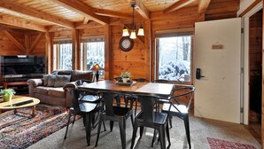 A cozy cabin interior with a wooden ceiling, a dining table with chairs, a sofa, a TV, and a view of snow-covered trees through large windows. - Killington Cabin: Right Unit - Sleeps 10 in 4 BR, 2 BA Cozy Escape (Killington)