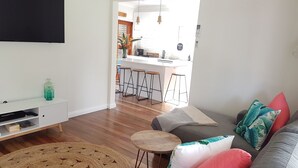 A modern living room with a sofa, a coffee table, and a TV stand. There is a kitchen area with a bar stool and a plant.