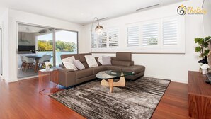 A modern living room with a brown sectional sofa, a glass coffee table, and a dining area with a table and chairs. - Luxury in Corlette - 4 Drungall Ave - Beautiful Home with Air Con and WiFi (CORLETTE)