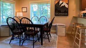 A dining area with a table and chairs, a window with a view of greenery, a wall-mounted picture, and a kitchen area with a refrigerator and bar stools. - Mountain Harbor #205 3 Bedroom Condo (Whitefish)