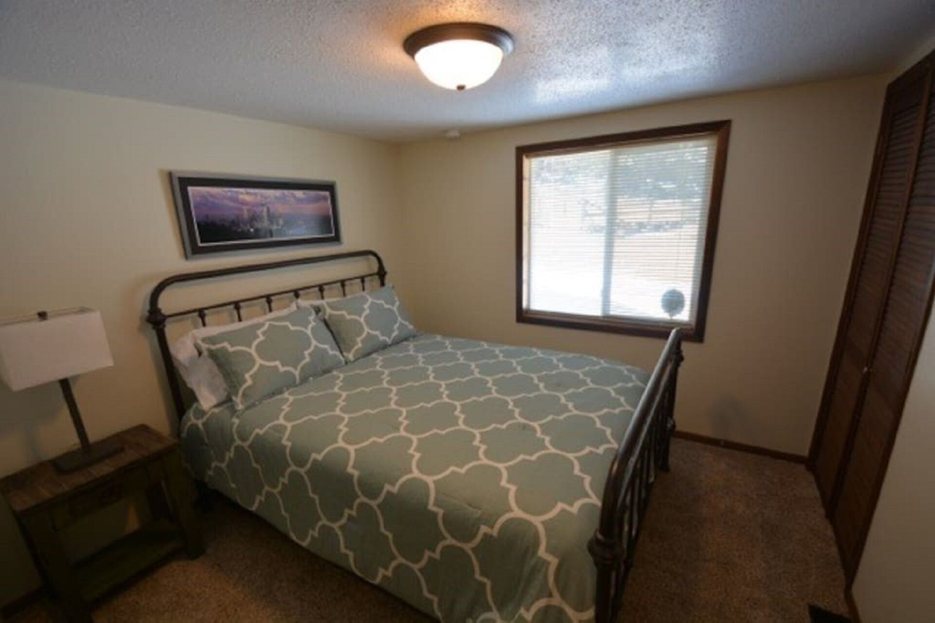 A bedroom with a bed, a nightstand, a lamp, a window with blinds, and a door.