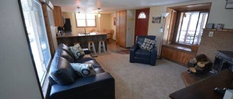 A living room with a sofa, a chair, a small table, and a fireplace. There is a kitchen area with a counter and stools.