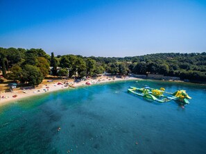 Une vue aérienne d’une plage aux eaux turquoise et limpides, avec un rivage sablonneux et quelques personnes qui se baignent.